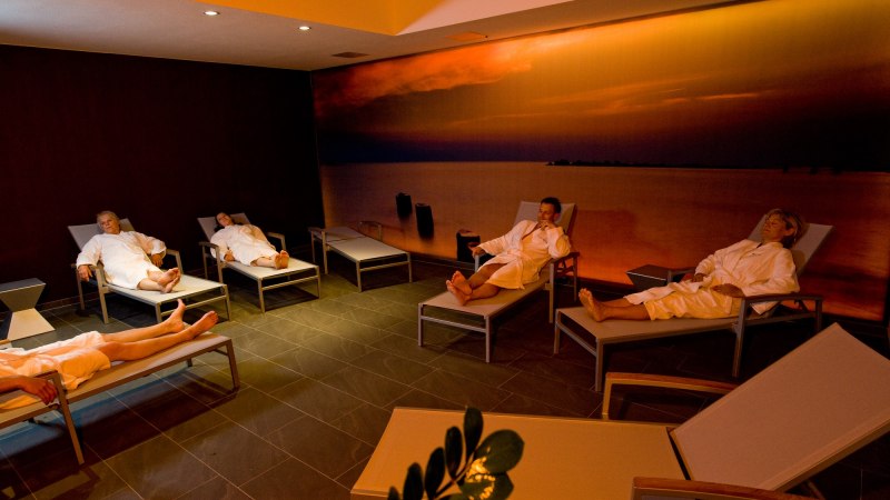 Personen in Bademänteln entspannen auf Liegen in einem Spa-Raum. Die Wand zeigt ein Bild eines Sonnenuntergangs über dem Meer., © Hotel Fortuna