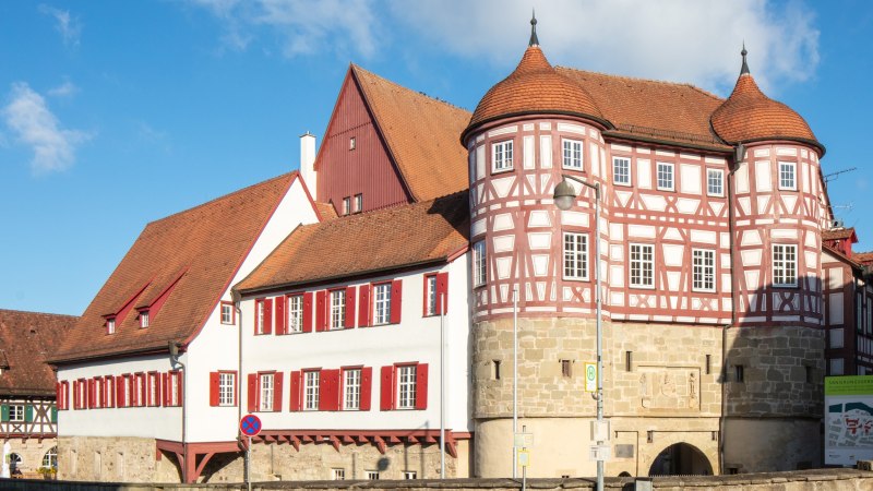 altes Schloss - rechts das Torhaus mit Sockelgeschoss aus hellem Sandstein und aufgesetztem roten Fachwerk mit weißen Gefachen, flankiert von zwei Rundtürmen. In der Bildmitte der Westflügel mit zwei aneinandergebauten, weiß verputzten Gebäudeteilen mit roten Fensterläden, überragt vom Dach und rotbraun gestrichener Giebelseite des Nordflügels. Alle Gebäudeteile eingedekct mit roten Ziegeln. Links das Fachwerkgebäude der ehemaligen Bentheimschen Forstverwaltung mit hellem Sandsteinsockel und grünen Fensterläden., © Stadt Gaildorf altes Schloss - rechts das Torhaus mit Sockelgeschoss aus hellem Sandstein und aufgesetztem roten Fachwerk mit weißen Gefachen, flankiert von zwei Rundtürmen. In der Bildmitte der Westflügel mit zwei aneinandergebauten, weiß verputzten Gebäudeteilen mit roten Fensterläden, überragt vom Dach und rotbraun gestrichener Giebelseite des Nordflügels. Alle Gebäudeteile eingedekct mit roten Ziegeln. Links das Fachwerkgebäude der ehemaligen Bentheimschen Forstverwaltung mit hellem Sandsteinsockel und grünen Fensterläden., © Stadt Gaildorf