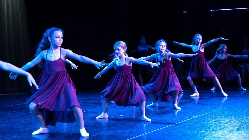 Kinder in lila Kleidern tanzen auf einer Bühne. Sie sind in einer synchronen Pose und tragen weiße Socken. Die Bühne ist dunkel beleuchtet., © Kunstschule Labyrinth