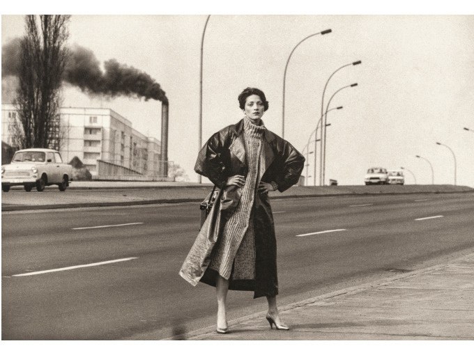 Eine Frau in einem langen Mantel posiert auf einer leeren Stra&szlig;e. Im Hintergrund sind rauchende Schornsteine und Autos zu sehen, was eine industrielle Umgebung andeutet., &copy; Kunsthalle G&ouml;ppingen