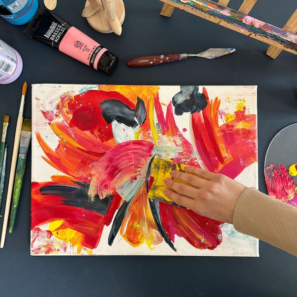 Hand malt mit Schwamm auf abstrakter Leinwand in Rot, Gelb und Schwarz. Pinsel und Farben liegen verstreut auf einem Tisch., © Mercedes-Benz AG