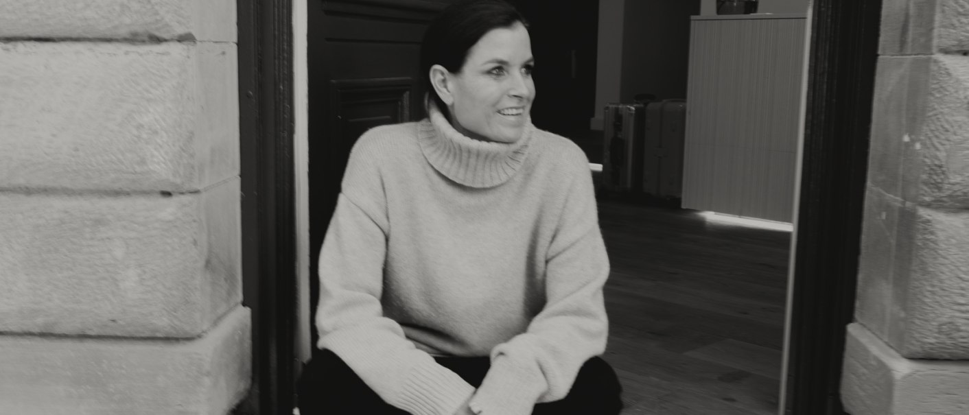 Eine Frau sitzt lächelnd auf einer Türschwelle. Sie trägt einen Rollkragenpullover und Sneakers. Der Hintergrund zeigt einen Innenraum., © Peter Oliver Wolff / DAS EDITH