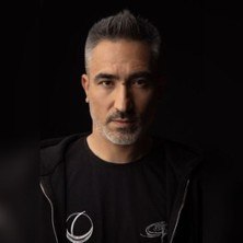 Ein Mann mit grauem Haar und Bart blickt ernst in die Kamera. Er tr&auml;gt ein schwarzes T-Shirt und eine schwarze Jacke. Der Hintergrund ist dunkel., &copy; links im Bild