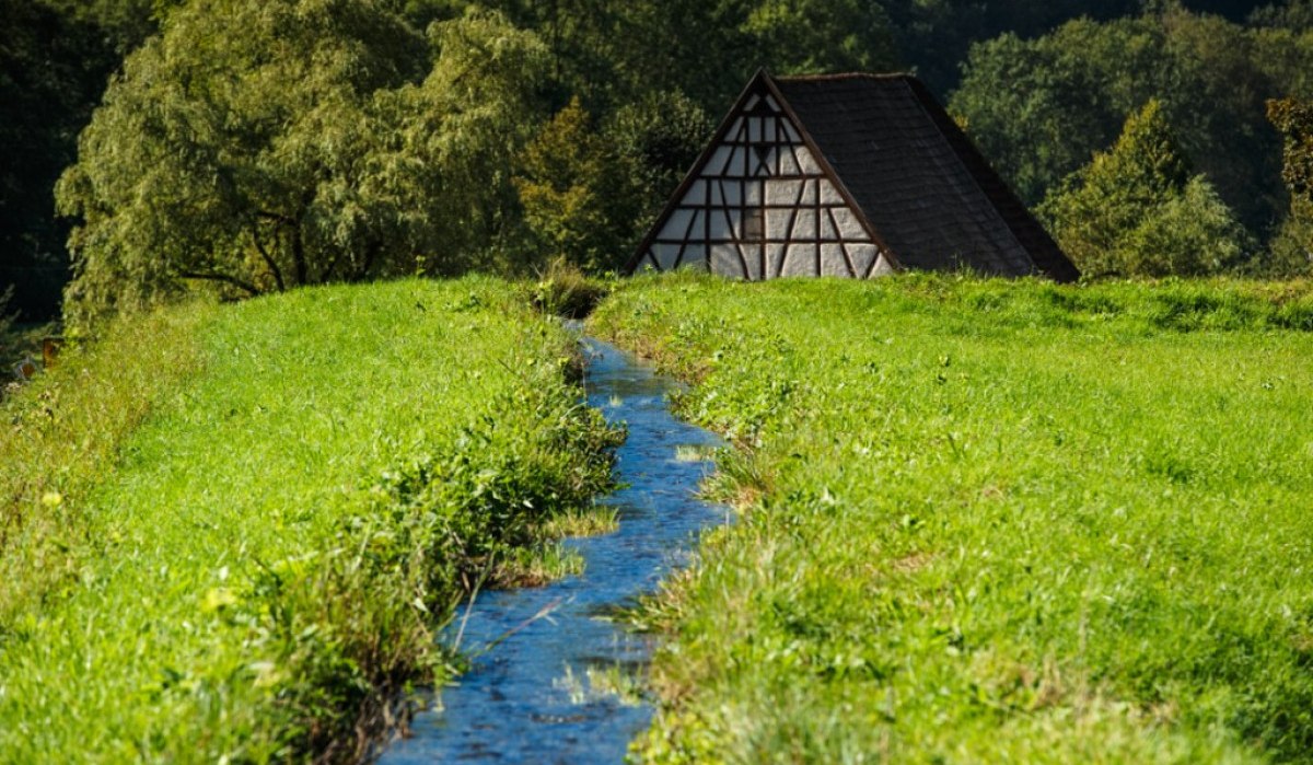 Ein kleines Fachwerkhaus steht im Eybtal, umgeben von grüner Wiese und einem schmalen Bach. Im Hintergrund sind Bäume zu sehen., © Landratsamt Göppingen Ein kleines Fachwerkhaus steht im Eybtal, umgeben von grüner Wiese und einem schmalen Bach. Im Hintergrund sind Bäume zu sehen., © Landratsamt Göppingen