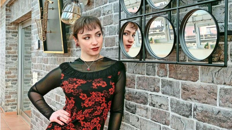 Eine Frau in einem schwarz-roten Kleid posiert vor einer Backsteinwand mit runden Spiegeln, die ihr Spiegelbild zeigen. Dekorative Elemente an der Wand., &copy; Foto Simon Granville