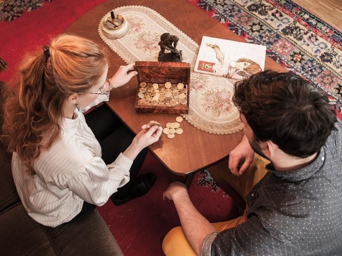 Zwei Personen sitzen an einem Tisch und betrachten eine Schachtel voller M&uuml;nzen. Auf dem Tisch liegt ein Tischl&auml;ufer und ein Buch., &copy; ExitGames Stuttgart