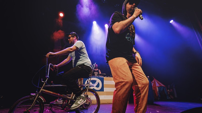 Ein Mann rappt auf der Bühne, während ein anderer auf einem Fahrrad fährt. Bunte Bühnenbeleuchtung und ein DJ-Pult im Hintergrund., © Scala Kultur Live gGmbH
