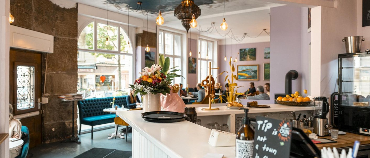 Gemütliches Café mit Blumen und Brezeln auf der Theke, blauen Sofas und Kunst an den Wänden. Große Fenster lassen viel Licht herein., © SMG, Sarah Schmid Gemütliches Café mit Blumen und Brezeln auf der Theke, blauen Sofas und Kunst an den Wänden. Große Fenster lassen viel Licht herein., © SMG, Sarah Schmid