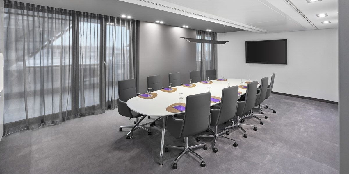 Moderner Konferenzraum mit ovalem Tisch, grauen Stühlen und großem Bildschirm an der Wand., © Jaz Hotel Stuttgart