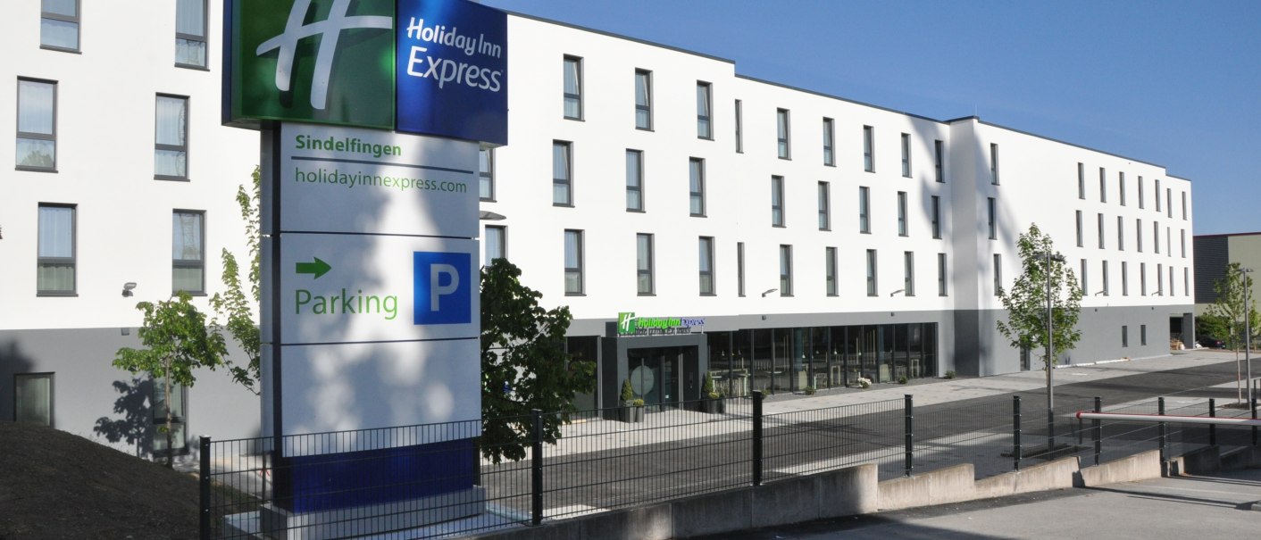 Das Holiday Inn Express in Sindelfingen zeigt eine moderne Fassade mit einem großen Parkplatzschild im Vordergrund. Der Himmel ist klar und blau., © tristar GmbH Das Holiday Inn Express in Sindelfingen zeigt eine moderne Fassade mit einem großen Parkplatzschild im Vordergrund. Der Himmel ist klar und blau., © tristar GmbH