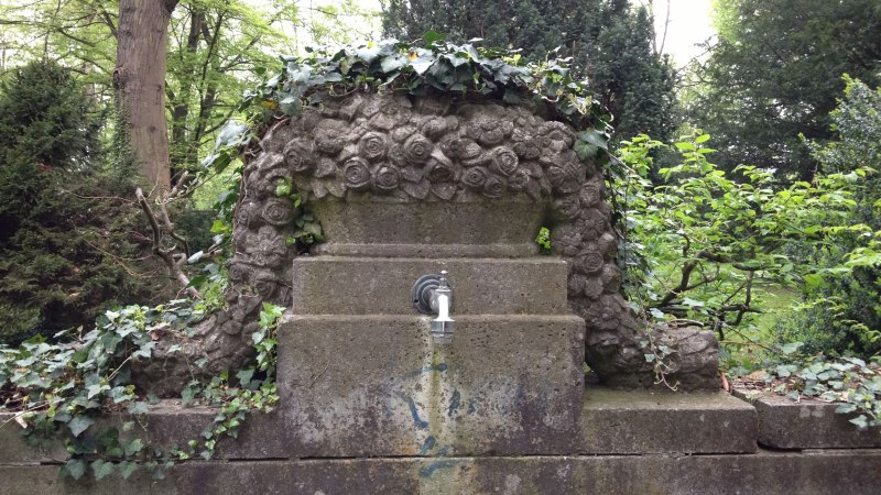 Ein alter Steinbrunnen mit Rosenverzierungen, umgeben von Efeu und B&auml;umen, steht auf einem Friedhof., &copy; StadtN&uuml;rtingen