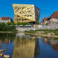 Ein modernes, goldenes Gebäude spiegelt sich in einem Fluss. Historische Gebäude und grüne Uferpflanzen umgeben die Szene unter klarem, blauem Himmel., © Touristik und Marketing GmbH
