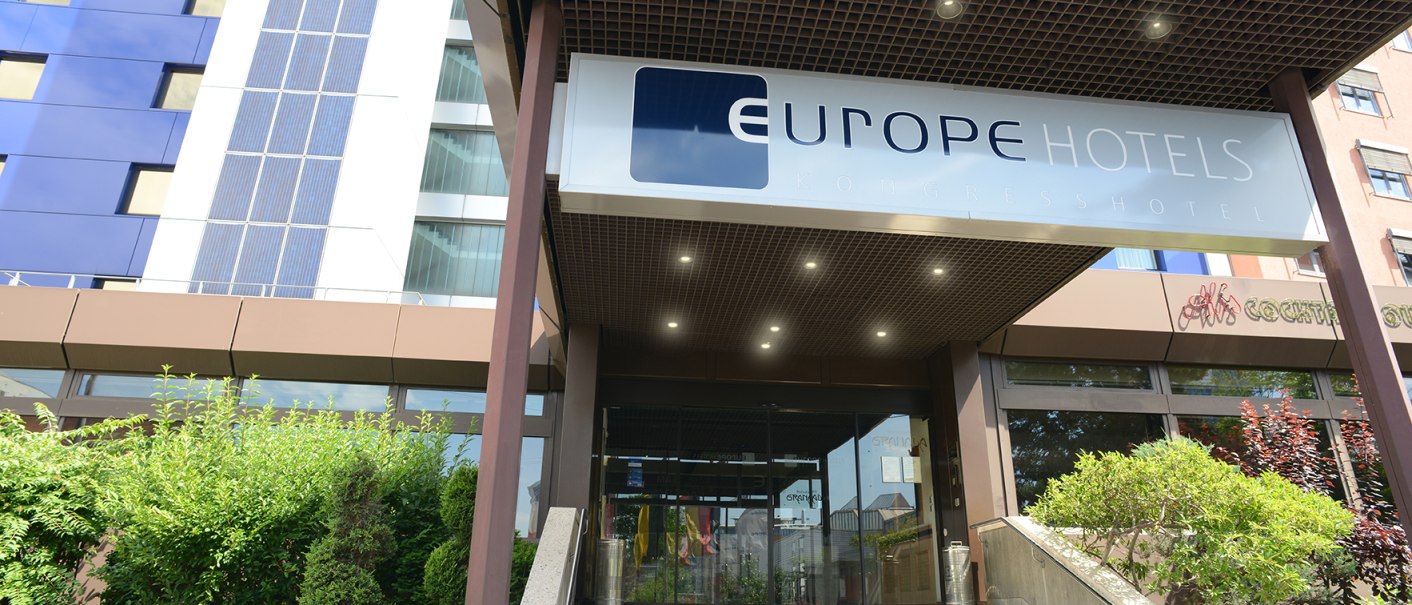 Eingang eines modernen Hotels mit dem Schild 'Europe Hotels Kongresshotel' über der Treppe. Links sind grüne Büsche zu sehen., © Europe Hotels International