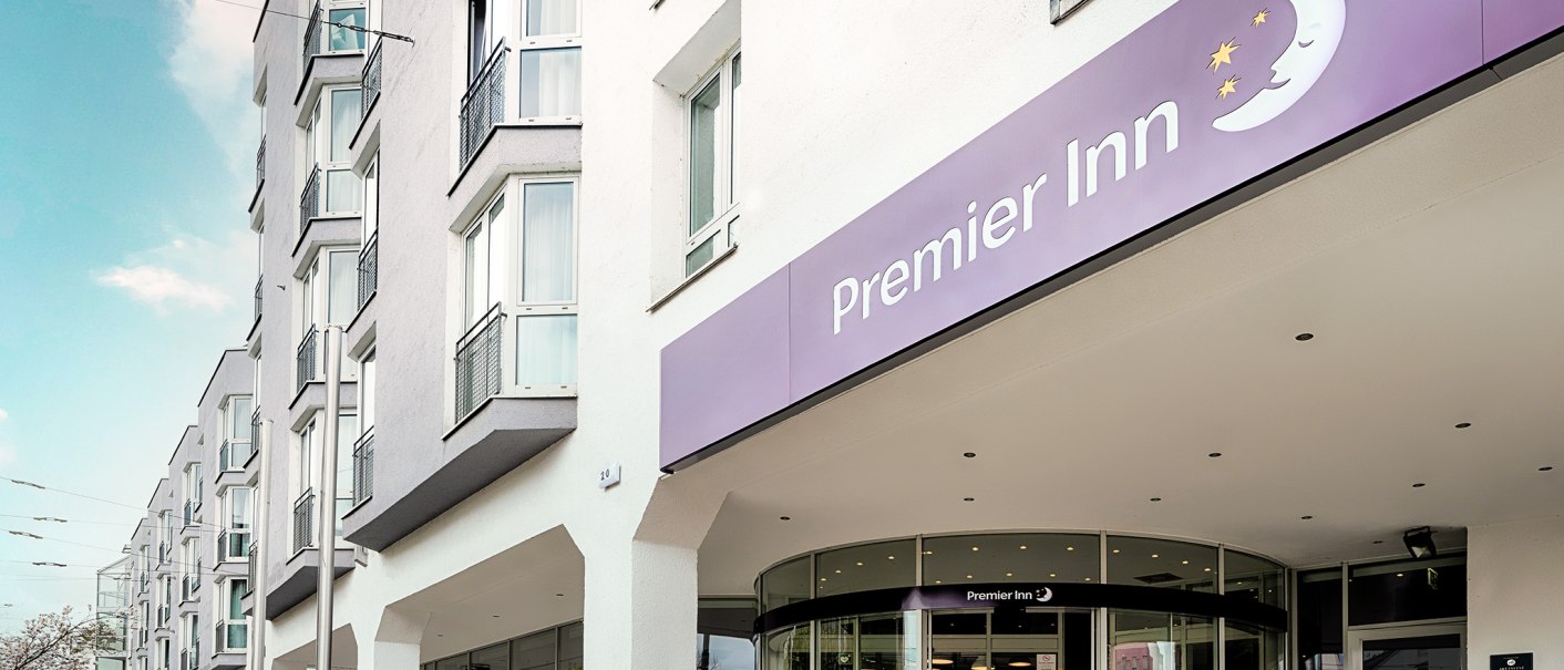 Außenansicht eines Premier Inn Hotels mit lila Schild und Eingangstür. Mehrstöckiges Gebäude mit großen Fenstern., © Premier Inn Germany