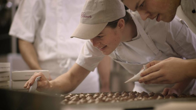 Zwei Personen in wei&szlig;en Sch&uuml;rzen dekorieren Pralinen in einer Konditorei. Sie konzentrieren sich auf die Details der Schokoladenkreationen.