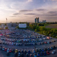 Luftaufnahme des Autokinos Kornwestheim bei Sonnenuntergang. Viele Autos sind geparkt, eine große Leinwand ist sichtbar. Im Hintergrund Felder und Gebäude., © DriveIn Autokinos DWJ GmbH