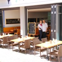 Ein Restaurant mit offenen Türen, draußen stehen Tische und Stühle. Zwei Personen stehen im Eingang neben einer Tafel mit Menü.