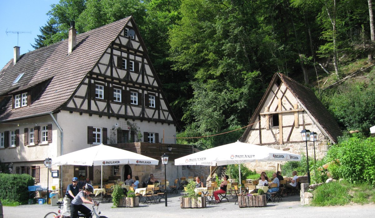 Ein Fachwerkhaus mit Biergarten, umgeben von Bäumen. Menschen sitzen an Tischen, während zwei Radfahrer im Vordergrund stehen., © Natur.Nah. Schönbuch & Heckengäu Ein Fachwerkhaus mit Biergarten, umgeben von Bäumen. Menschen sitzen an Tischen, während zwei Radfahrer im Vordergrund stehen., © Natur.Nah. Schönbuch & Heckengäu