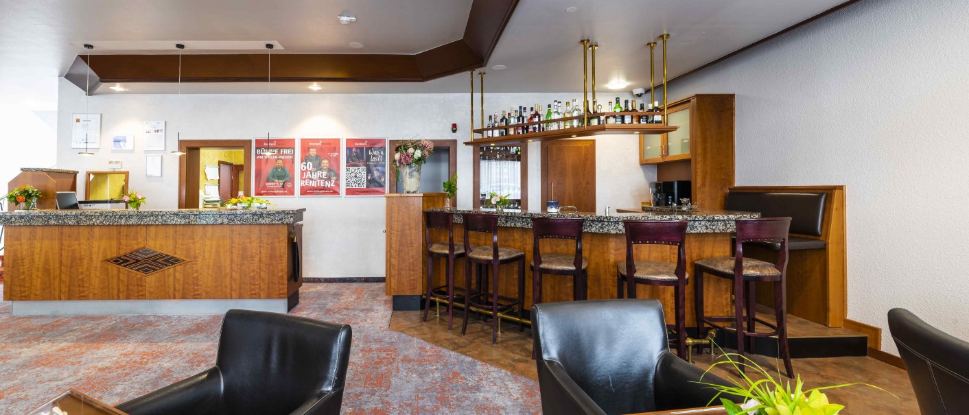 Hotelbar mit Holzmöbeln, Barhockern und Dekorationen. An der Wand hängen Poster, auf den Tischen stehen Blumen., © TOMAS