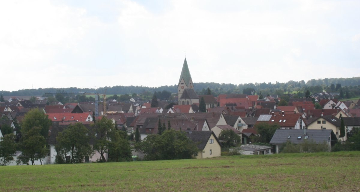 Dorfansicht mit Kirche und roten Dächern, umgeben von Bäumen und Feldern unter bewölktem Himmel., © Natur.Nah. Schönbuch & Heckengäu Dorfansicht mit Kirche und roten Dächern, umgeben von Bäumen und Feldern unter bewölktem Himmel., © Natur.Nah. Schönbuch & Heckengäu