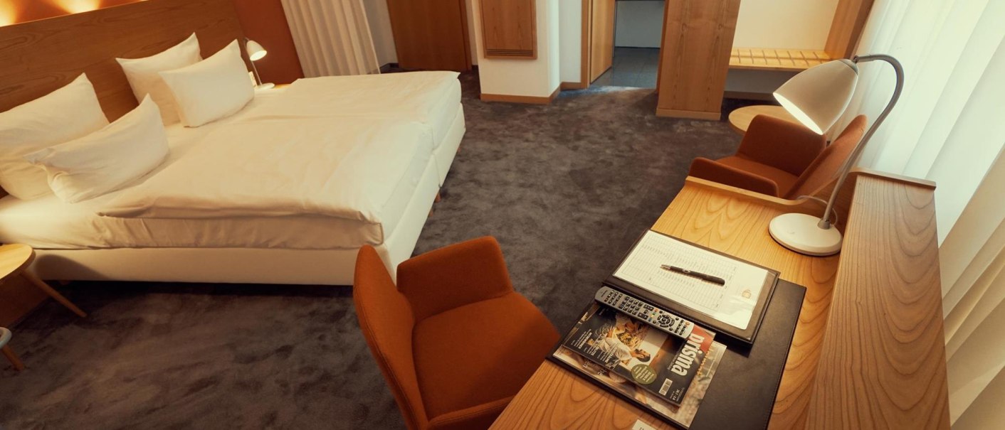 Modernes Hotelzimmer mit Doppelbett, Schreibtisch und Stuhl. Eine Lampe beleuchtet den Arbeitsbereich. Teppichboden und warme Farben dominieren den Raum., © Ringhotel Adler Asperg
