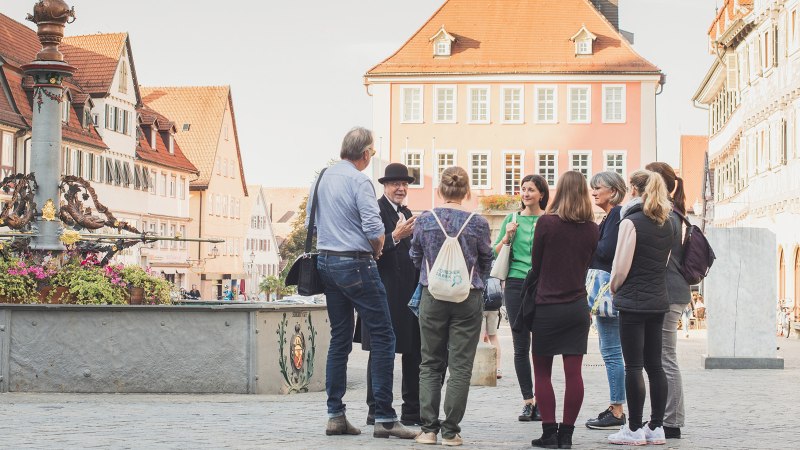 Eine Gruppe von Menschen steht um einen Marktbrunnen in einer Altstadt mit Fachwerkh&auml;usern. Ein Mann in Hut und Mantel scheint eine F&uuml;hrung zu geben., &copy; Bebop Media, Danijel Grbic
