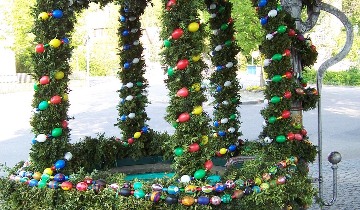 Ein traditionell geschmückter Osterbrunnen mit bunten Eiern und grünen Girlanden, umgeben von Bäumen und gepflastertem Boden., © Natur.Nah. Schönbuch & Heckengäu Ein traditionell geschmückter Osterbrunnen mit bunten Eiern und grünen Girlanden, umgeben von Bäumen und gepflastertem Boden., © Natur.Nah. Schönbuch & Heckengäu