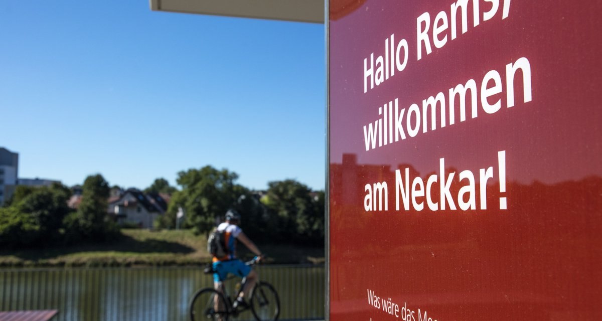 Ein Schild mit der Aufschrift 'Hallo Rems, willkommen am Neckar!' im Vordergrund, ein Radfahrer fährt am Fluss entlang. Ein Schild mit der Aufschrift 'Hallo Rems, willkommen am Neckar!' im Vordergrund, ein Radfahrer fährt am Fluss entlang.