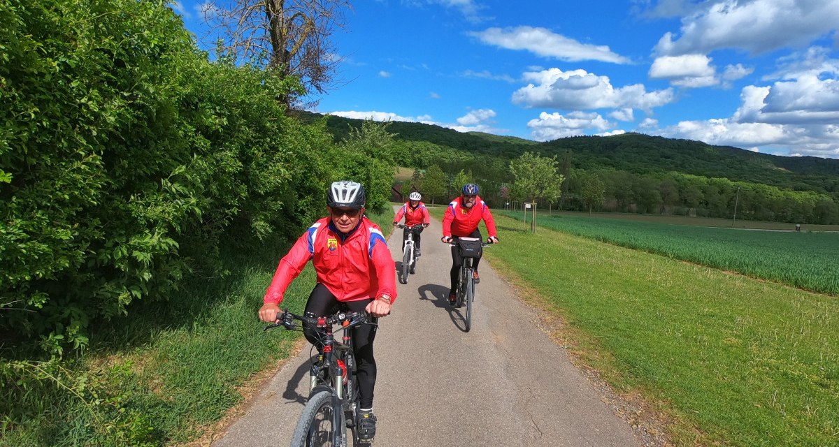 Drei Radfahrer in roten Jacken fahren auf einem ländlichen Weg. Die Umgebung ist grün, mit Bäumen und Feldern, unter einem blauen Himmel mit Wolken., © Land der 1000 Hügel - Kraichgau-Stromberg