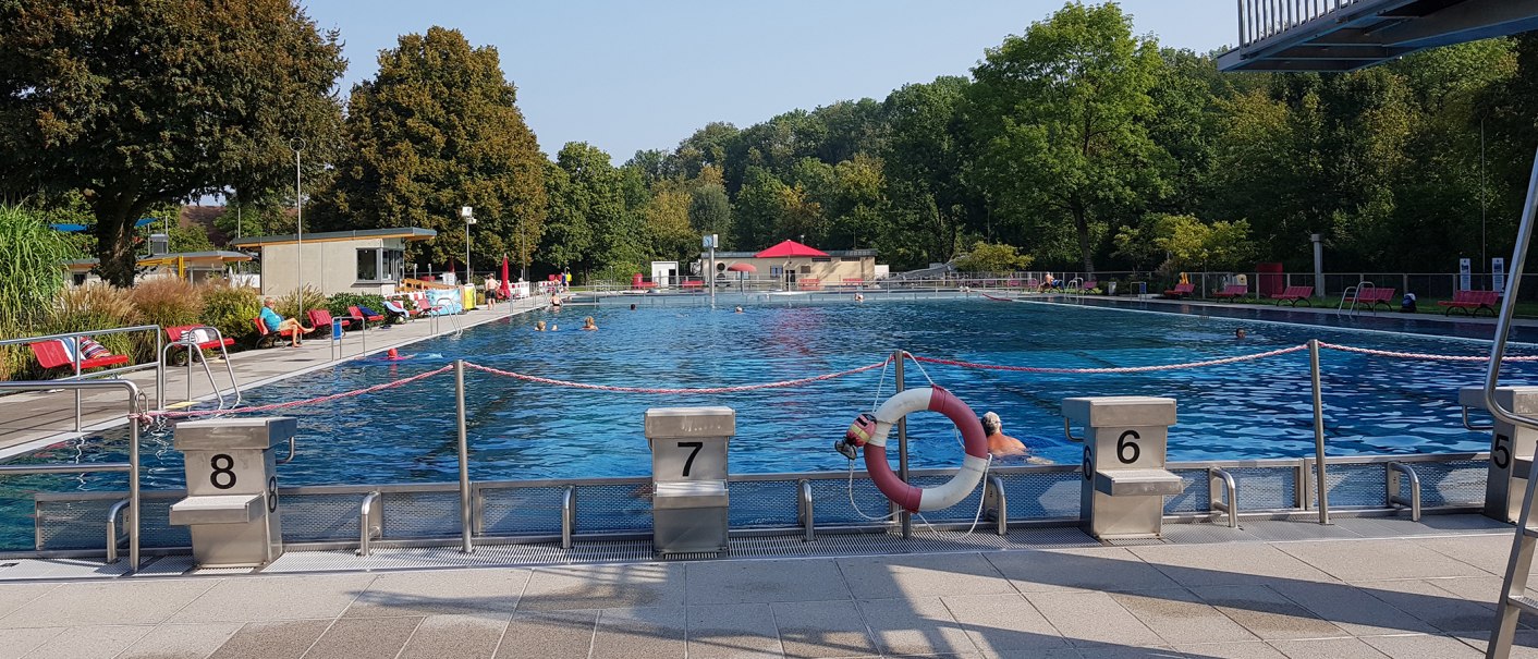 Ein Freibad mit mehreren Schwimmern, umgeben von Bäumen. Im Vordergrund sind Startblöcke und ein Rettungsring zu sehen., © Torsten Wenzler
