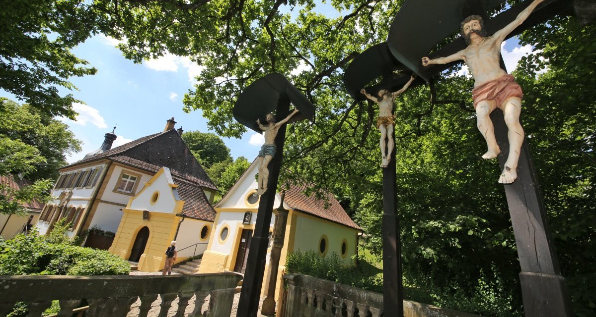 Drei aus Holz geschnitzte Personen hängen an Kreuzen. Im Hintergrund stehen drei Häuser., © Foto: Walter Laible Drei aus Holz geschnitzte Personen hängen an Kreuzen. Im Hintergrund stehen drei Häuser., © Foto: Walter Laible