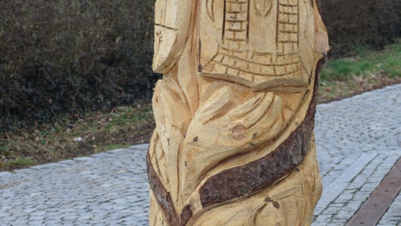 Holzskulptur mit geschnitzten Motiven, darunter ein Gesicht und Geb&auml;ude, auf einem gepflasterten Weg am Skulpturenpfad B&ouml;bingen., &copy; Gemeinde B&ouml;bingen