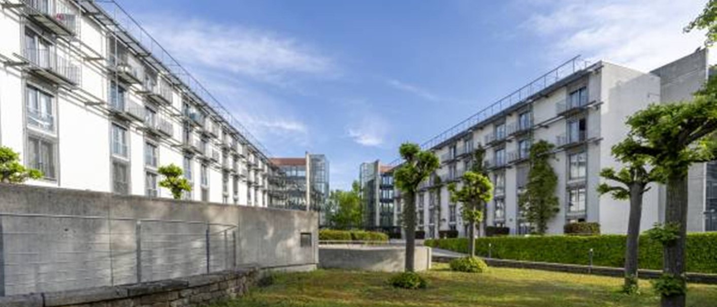 Zwei moderne Gebäude mit Glasfassaden und Balkonen, dazwischen ein gepflegter Garten mit Bäumen, unter blauem Himmel., © TOMAS Zwei moderne Gebäude mit Glasfassaden und Balkonen, dazwischen ein gepflegter Garten mit Bäumen, unter blauem Himmel., © TOMAS
