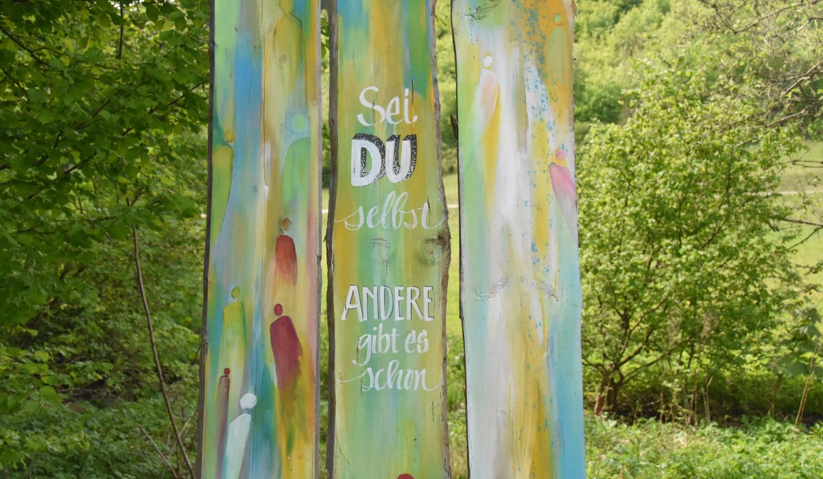 Bunte Holzstele im Wald mit dem Spruch 'Sei du selbst, andere gibt es schon'. Umgeben von grüner Natur., © Ev. Kirchenbezirk Schorndorf