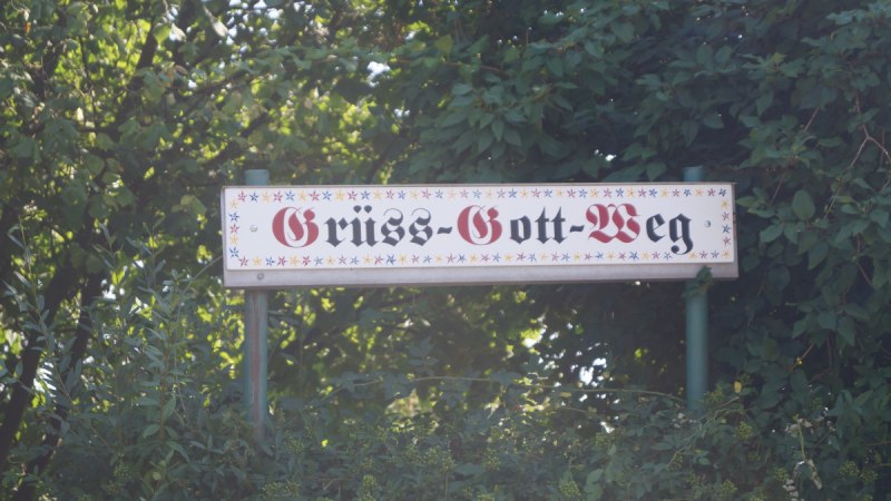 Ein kalligraphisch gestaltetes Schild mit der Aufschrift 'Grüß-Gott-Weg', umgeben von grünen Bäumen und Pflanzen., © Ludwigsburg - Stuttgart-Marketing GmbH Ein kalligraphisch gestaltetes Schild mit der Aufschrift 'Grüß-Gott-Weg', umgeben von grünen Bäumen und Pflanzen., © Ludwigsburg - Stuttgart-Marketing GmbH