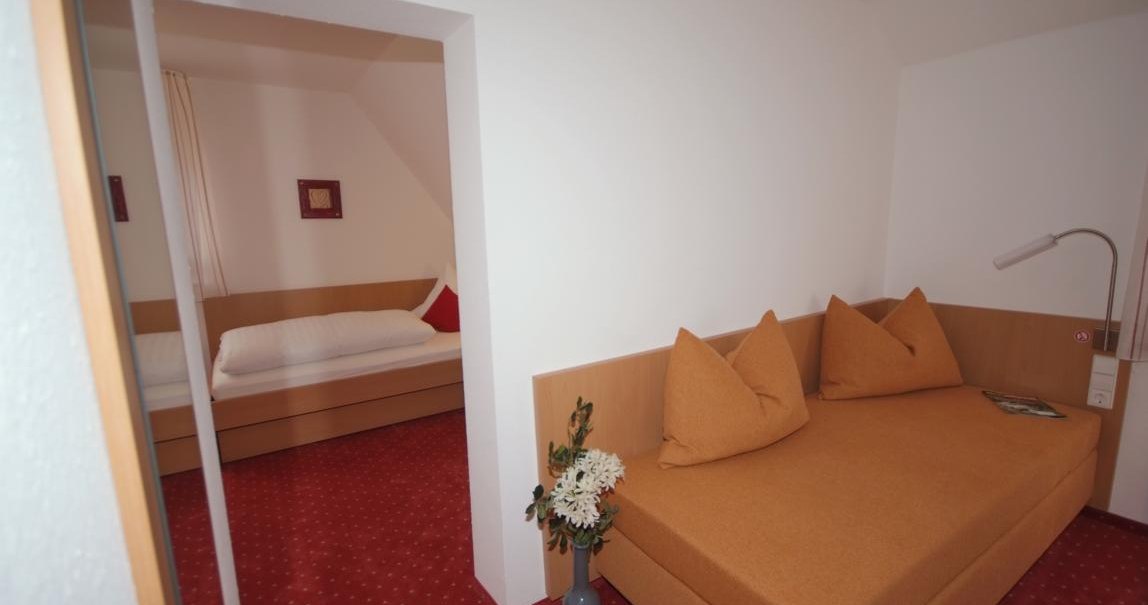 Kleines Zimmer mit Einzelbett und Sofa, roter Teppichboden, weiße Wände. Vase mit Blumen auf dem Boden, Lampe und Kissen auf dem Sofa., © TOMAS