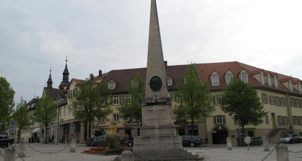 Ein Obelisk steht auf einem gepflasterten Platz, umgeben von historischen Gebäuden und Bäumen. Der Himmel ist bewölkt., © Ludwigsburg - Stuttgart-Marketing GmbH Ein Obelisk steht auf einem gepflasterten Platz, umgeben von historischen Gebäuden und Bäumen. Der Himmel ist bewölkt., © Ludwigsburg - Stuttgart-Marketing GmbH
