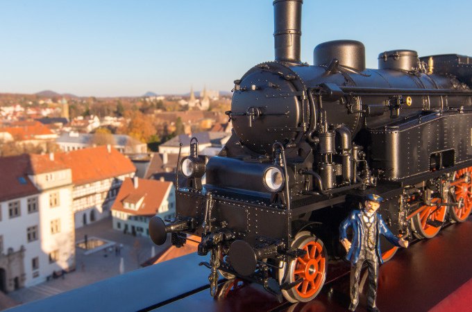 Eine schwarze Modelllokomotive und eine Lokführerfigur stehen auf einem Geländer. Im Hintergrund ist eine Stadtlandschaft bei klarem Himmel zu sehen., © Stadt Göppingen Eine schwarze Modelllokomotive und eine Lokführerfigur stehen auf einem Geländer. Im Hintergrund ist eine Stadtlandschaft bei klarem Himmel zu sehen., © Stadt Göppingen