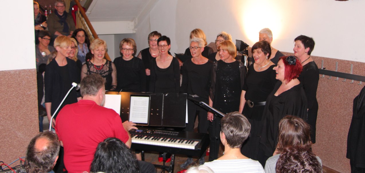 Ein Chor von Frauen singt bei einer Veranstaltung, begleitet von einem Mann am Klavier. Zuschauer sitzen und stehen im Hintergrund., &copy; TonArt