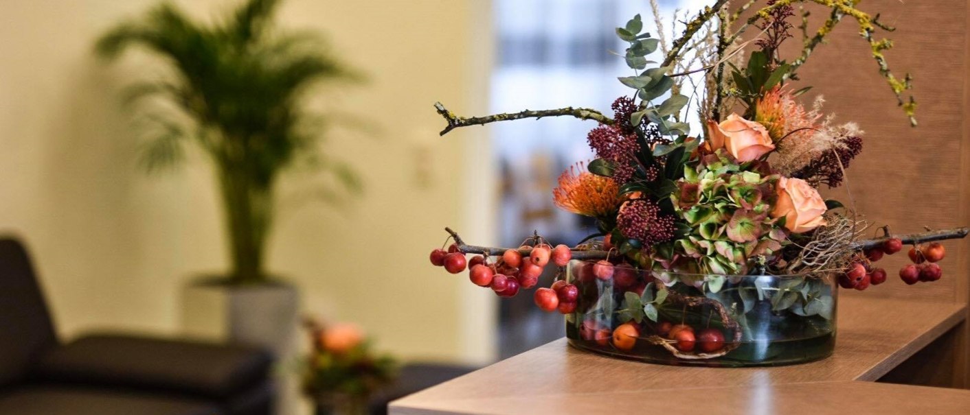 Blumenarrangement in Glasvase auf Empfangstresen, im Hintergrund Couch und Pflanze., © TOMAS Blumenarrangement in Glasvase auf Empfangstresen, im Hintergrund Couch und Pflanze., © TOMAS