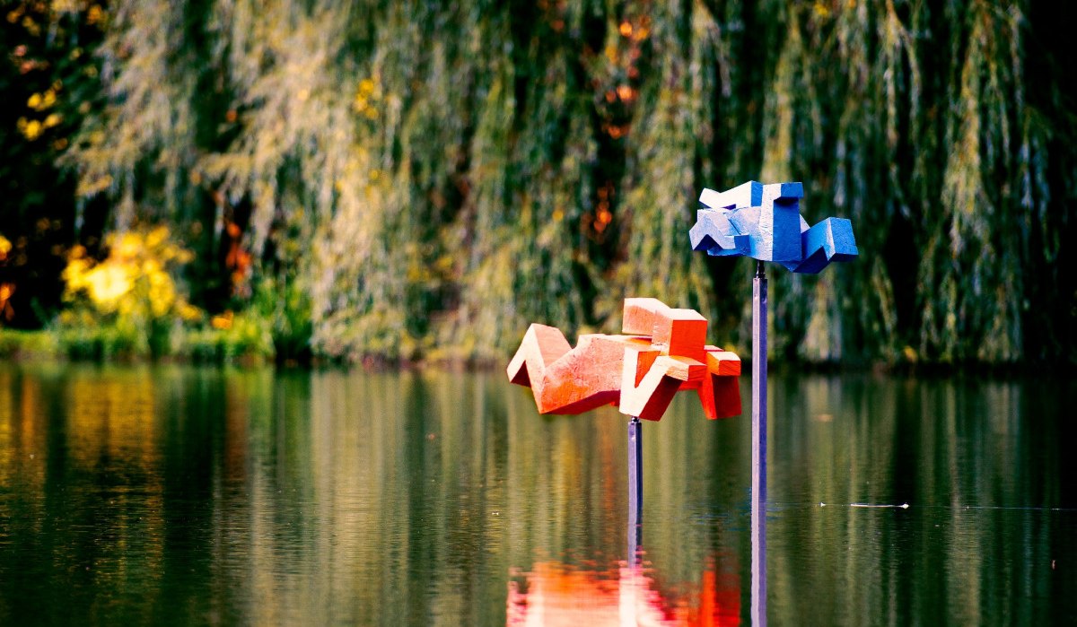 Zwei geometrische Skulpturen in Blau und Rot, die Schwimmer darstellen, stehen auf Stangen im Wasser eines Sees, umgeben von üppiger Vegetation., © Natur.Nah. Schönbuch & Heckengäu Zwei geometrische Skulpturen in Blau und Rot, die Schwimmer darstellen, stehen auf Stangen im Wasser eines Sees, umgeben von üppiger Vegetation., © Natur.Nah. Schönbuch & Heckengäu