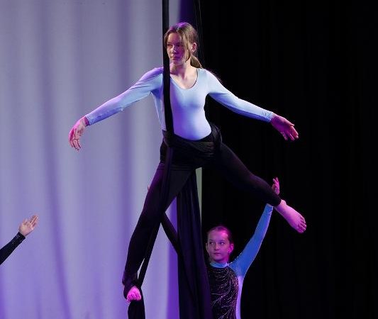 Ein Mädchen in einem blauen Trikot führt Luftakrobatik an schwarzen Tüchern auf einer Bühne aus. Ein weiteres Kind assistiert ihr., © Kunstschule Labyrinth Ein Mädchen in einem blauen Trikot führt Luftakrobatik an schwarzen Tüchern auf einer Bühne aus. Ein weiteres Kind assistiert ihr., © Kunstschule Labyrinth