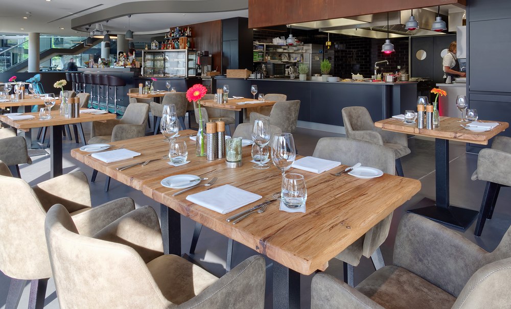 Modernes Restaurant mit Holztischen, gedeckt mit Gläsern und Besteck. Im Hintergrund eine offene Küche und Bar., © V8 HOTEL Motorworld Region Stuttgart Modernes Restaurant mit Holztischen, gedeckt mit Gläsern und Besteck. Im Hintergrund eine offene Küche und Bar., © V8 HOTEL Motorworld Region Stuttgart