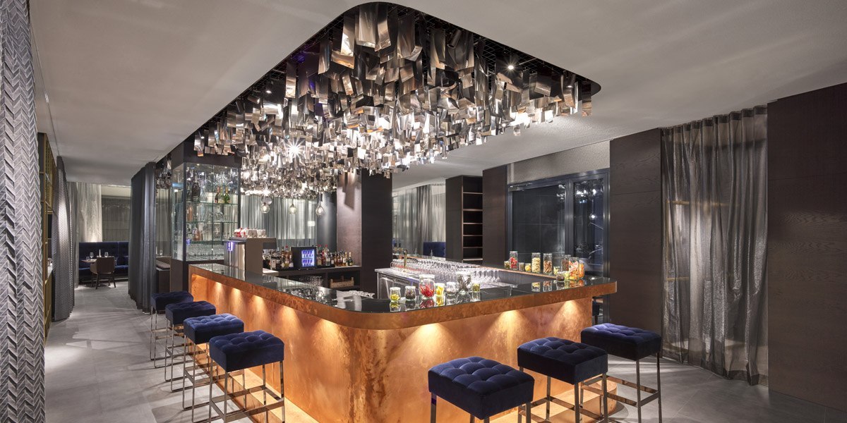 Elegante Bar mit beleuchtetem Tresen, blauen Hockern und einer auffälligen Deckeninstallation aus Metall. Stilvolle, moderne Atmosphäre., © Jaz Hotel Stuttgart