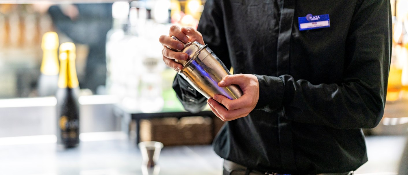 Ein Barkeeper in schwarzer Kleidung hält einen Cocktailshaker in der Hand. Im Hintergrund sind Flaschen und Barzubehör zu sehen., © Plaza Residence Stuttgart-Ditzingen Ein Barkeeper in schwarzer Kleidung hält einen Cocktailshaker in der Hand. Im Hintergrund sind Flaschen und Barzubehör zu sehen., © Plaza Residence Stuttgart-Ditzingen
