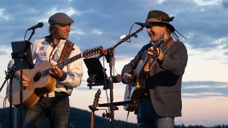 Zwei Musiker spielen Gitarren bei einem Open-Air-Konzert. Der Himmel ist bewölkt, und die Sonne geht unter. Beide tragen Hüte und lächeln., © Backnang Zwei Musiker spielen Gitarren bei einem Open-Air-Konzert. Der Himmel ist bewölkt, und die Sonne geht unter. Beide tragen Hüte und lächeln., © Backnang