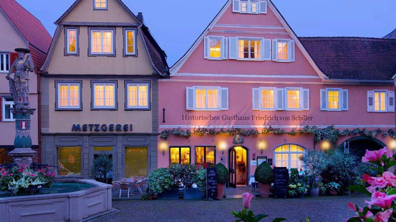 Ein malerisches Dorf bei Dämmerung mit einem historischen Gasthaus und einer Metzgerei. Blumen und ein Brunnen schmücken den Platz.