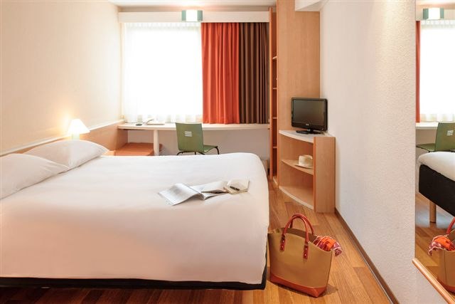 Helles Hotelzimmer mit Doppelbett, Schreibtisch, Fernseher und Tasche auf dem Boden. Vorhänge in Rot- und Brauntönen., © TOMAS