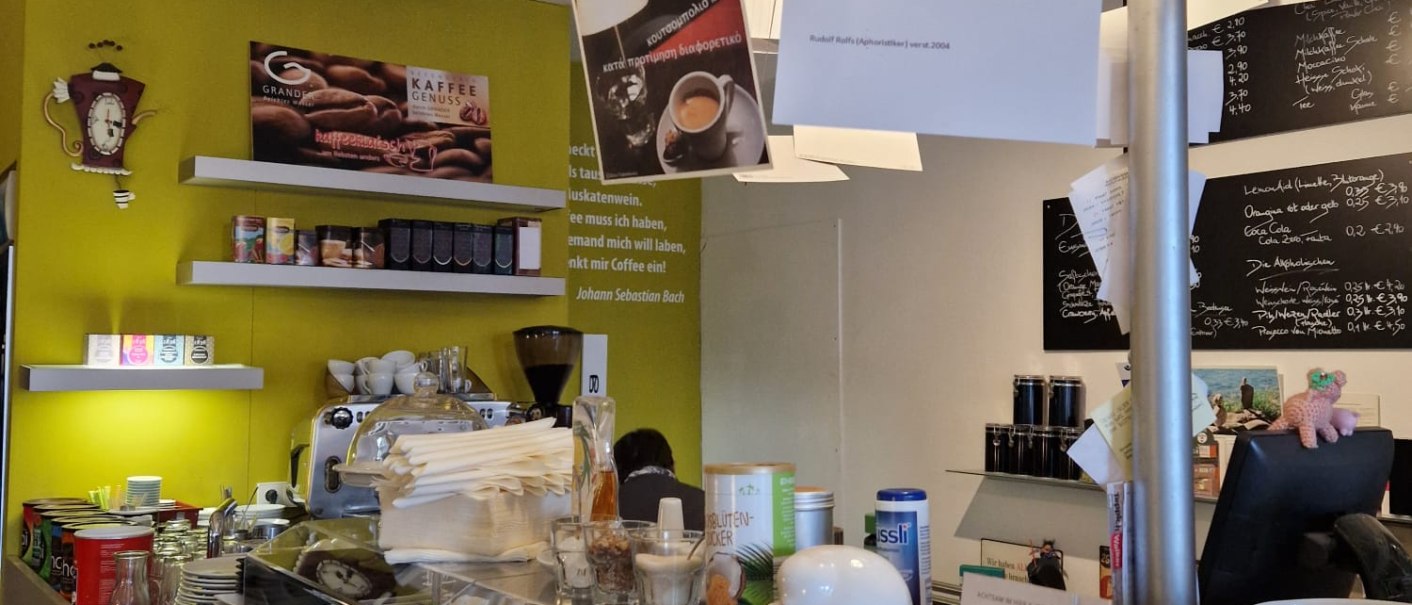 Ein Café mit einer Theke voller Tassen, einer Kaffeemaschine und hängenden Schildern. Broschüren und Flyer sind auf der Theke ausgelegt., © cStadtNuertingen Ein Café mit einer Theke voller Tassen, einer Kaffeemaschine und hängenden Schildern. Broschüren und Flyer sind auf der Theke ausgelegt., © cStadtNuertingen