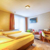 Ein gem&uuml;tliches Hotelzimmer mit Doppelbett, Schreibtisch, Fernseher und zwei Sesseln. Helle Vorh&auml;nge und bunte Kissen sorgen f&uuml;r eine einladende Atmosph&auml;re., &copy; TOMAS